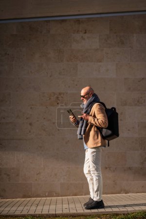 Photo for Mature businessman using mobile phone in the city. Man in casual clothes walking outdoors by a building. - Royalty Free Image