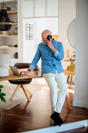Téléchargez les photos : Full length of happy middle aged man making a call and using laptop while working from home. Confident male standing at desk in the living room. Home office. - en image libre de droit