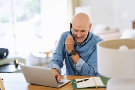 Téléchargez les photos : Happy middle aged man making a call and using laptop while working from home. Confident male sitting in the living room and having a call. Home office. - en image libre de droit