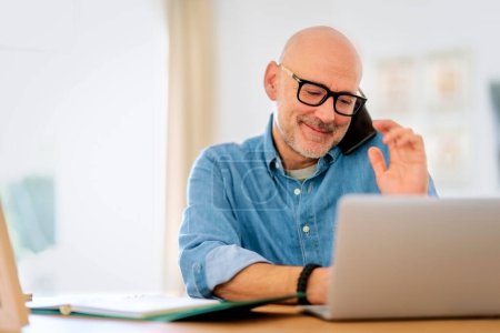 Photo for Happy middle aged man making a call and using laptop while working from home. Confident male sitting in the living room and having a call. Home office. - Royalty Free Image