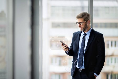 Foto de Confident businessman using smartphone and earphones while standing at the office. Professional man wearing suit and tie. - Imagen libre de derechos
