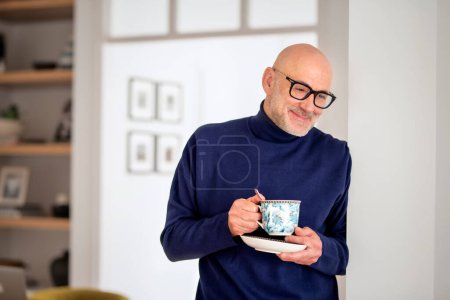 Foto de Middle aged man standing at the kitchen and daydreaming. Confident male wearing turtleneck sweater and drinking tea. - Imagen libre de derechos