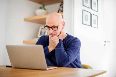 Photo for Thinking middle aged man using laptop and having video call while working from home. Confident male sitting at table at the kitchen. Home office. - Royalty Free Image