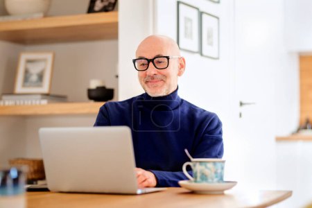 Téléchargez les photos : Confident middle aged man using laptop and having video call while working from home. Confident male sitting at table at the kitchen. Home office. - en image libre de droit
