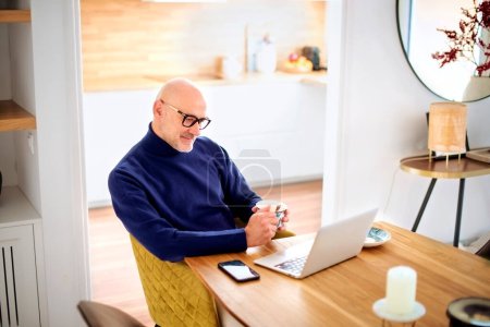 Téléchargez les photos : Confident middle aged man using laptop and having video call while working from home. Confident male drinking tea while sitting at table at the kitchen. Home office. - en image libre de droit