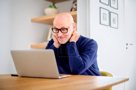 Téléchargez les photos : Thinking middle aged man using laptop and having video call while working from home. Confident male sitting at table at the kitchen. Home office. - en image libre de droit