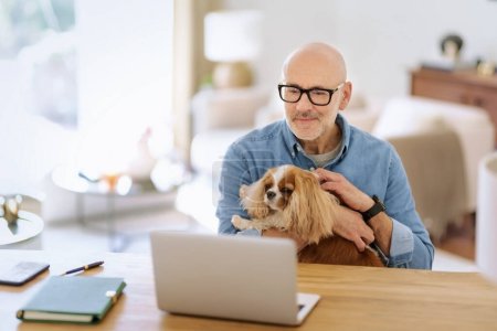 Téléchargez les photos : Thinking middle aged man using laptop and having video call while working from home. Confident male sitting at table with her cute puppy.   Home office. - en image libre de droit