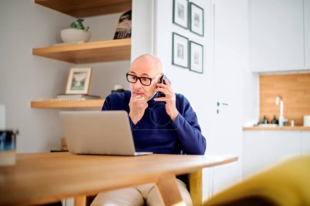 Téléchargez les photos : Wondered middle aged man making a call and using laptop while working from home. Confident male sitting in the dining room and having a call. Home office. - en image libre de droit