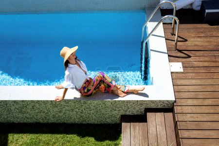 Foto de Happy woman wearing white shirt and sun hat while relaxing by the poolside. Full length shot. Copy space. - Imagen libre de derechos