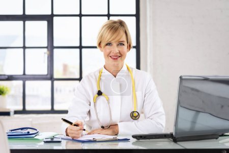 Téléchargez les photos : Smiling female doctor using laptop and writing something while sitting at doctors office. - en image libre de droit