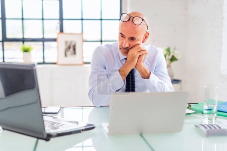 Foto de Hombre de negocios de mediana edad sosteniendo la cabeza y reflexionando mientras está sentado en la oficina y utilizando el ordenador portátil para el trabajo. - Imagen libre de derechos