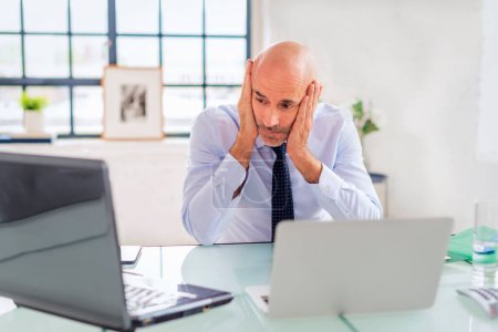 Téléchargez les photos : Homme d'affaires d'âge moyen tenant la tête et réfléchissant tout en étant assis au bureau et en utilisant un ordinateur portable pour le travail. - en image libre de droit