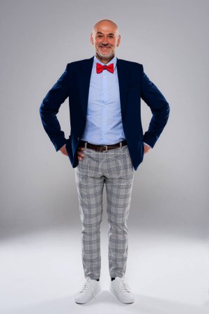 Foto de Full length studio shot of smiling man wearing bow tie and checked trousers while standing at isolated grey background. Copy space. - Imagen libre de derechos