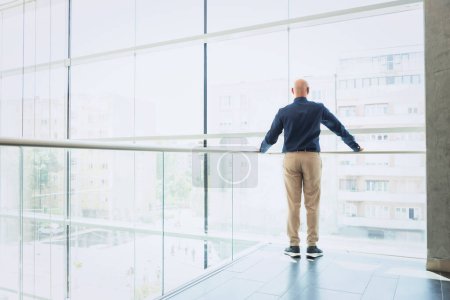 Photo for Rear view of thinking businessman looking out of office over city. Confident man wearing glasses and business casual. - Royalty Free Image