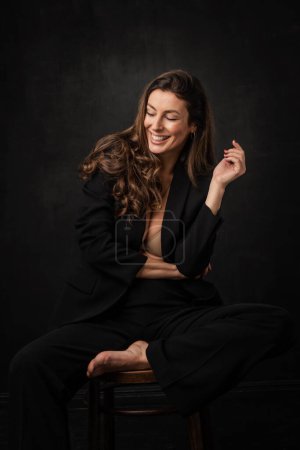 Photo for Full length of an attractive mid aged woman with toothy smile wearing black blazer and pants and sitting against at isolated dark background. Copy space. - Royalty Free Image