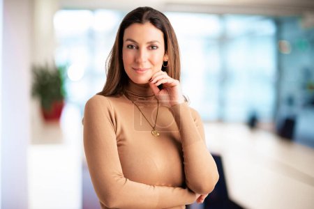 Photo for Close-up of an attractive middle aged female smiling and looking at camera. Brunette haired business woman wearing sweater while standing at the office. Copy space. - Royalty Free Image