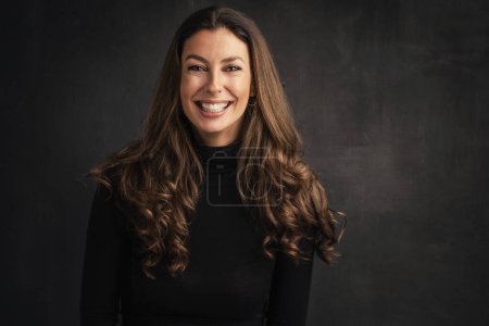 Foto de Headshot of an attractive middle aged woman with toothy smile wearing turtleneck sweater while sitting at isolated dark background. Copy space. Studio shot. Hand on forehead. - Imagen libre de derechos