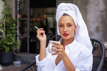 Foto de Attractive girl with towel turban on head wearing white shirt while having coffee on balcony and smoking cigarette in the morning. - Imagen libre de derechos