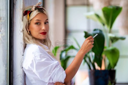 Foto de Portrait shot of beautiful young woman wearing hair scarf and white shirt while smoking cigarette at home. - Imagen libre de derechos
