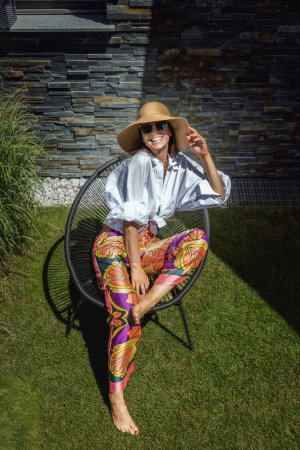 Foto de High angle shot of attractive woman wearing white shirt and straw hat while relaxing in a lounge chair in the garden. - Imagen libre de derechos