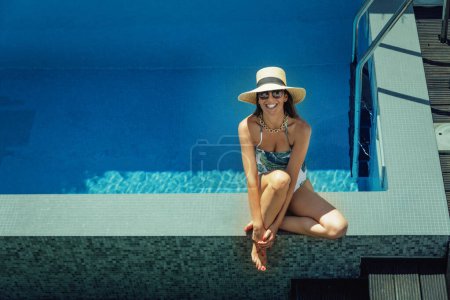Téléchargez les photos : Full length of a happy woman wearing swimsuit and straw hat while relaxing at the poolside. - en image libre de droit