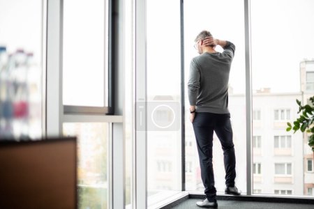 Photo for Rear view of thinking businessman looking out of office over city. Confident man wearing business casual. - Royalty Free Image