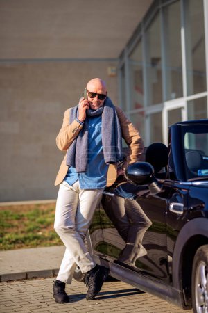 Téléchargez les photos : Plan complet de l'homme d'âge moyen portant des vêtements décontractés et utilisant un téléphone portable tout en restant debout à côté de sa voiture. - en image libre de droit