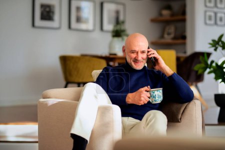 Photo for Portrait of middle aged man using cellphone and having a call while relaxing in an armchair at home. Happy male wearing turtleneck sweater and drinking tea. - Royalty Free Image