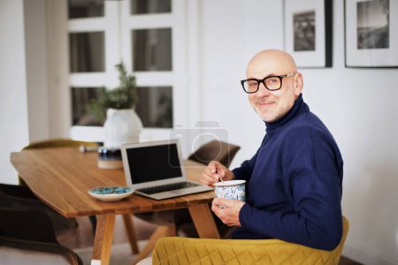 Foto de Retrato de un hombre de mediana edad que se relaja en una silla en casa y bebe té. Suéter y gafas de cuello alto para hombre feliz. - Imagen libre de derechos
