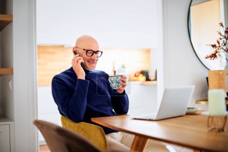 Photo for Middle aged man using laptop and smartphone while working from home. Confident male sitting at desk at home. Home office. - Royalty Free Image