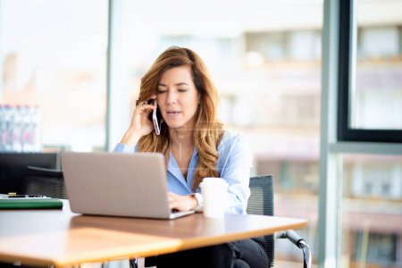 Photo for Candid shot of a mid aged professional woman wearing shirt and using laptop while working in a modern office. Attractive businesswoman sitting at desk and having a business call. - Royalty Free Image