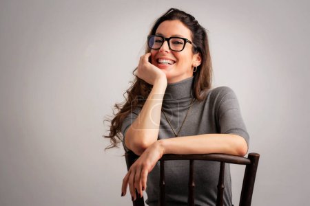 Photo for Close-up of an attractive middle aged woman with toothy smile wearing turtleneck sweater and eyewear while sitting at isolated grey background. Copy space. Studio shot. Hand on head. - Royalty Free Image