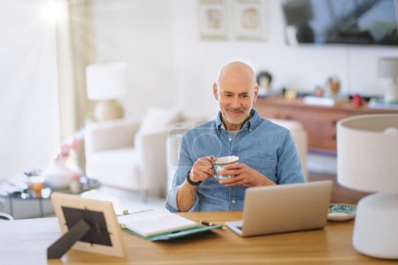 Photo for Confident middle aged man using laptop and having video call while working from home. Confident male drinking tea while sitting at table at the kitchen. Home office. - Royalty Free Image