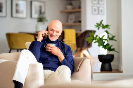 Photo for Mid aged man sitting in an armchair in a modern home and using smartphone. Confident man wearing casual clothes and drinking tea. - Royalty Free Image