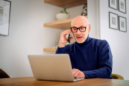 Photo for Surprised mid aged man making a call and using laptop while working from home. Confident male sitting in the living room and having a call. Home office. - Royalty Free Image
