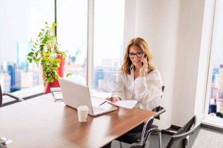 Téléchargez les photos : Femme d'affaires d'âge moyen assise dans un bureau moderne et ayant un appel d'affaires. Femme professionnelle confiante utilisant un ordinateur portable pour le travail. - en image libre de droit