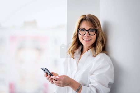 Photo for Confident businesswoman holding smart phone. Female executive is standing in a glass office and text messaging. She is wearing white shirt at office. - Royalty Free Image