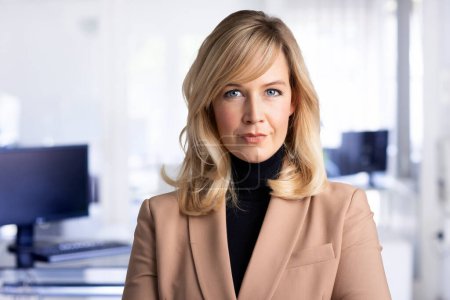 Photo for Close-up of mid aged businesswoman smiling and looking at camera. Blond haired female wearing blazer while standing at the office. Copy space. - Royalty Free Image