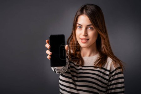 Photo for Attractive young woman presenting smart phone with blank screen. Caucasian female standing at isolated dark background. Copy space. - Royalty Free Image