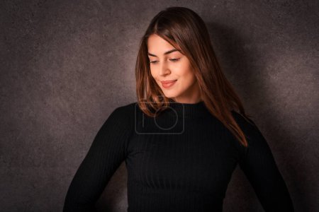 Photo for Portrait of smiling young woman standing at isolated background. Confident young female is having brunette hair. She is in black sweater against dark background. Copy space. - Royalty Free Image