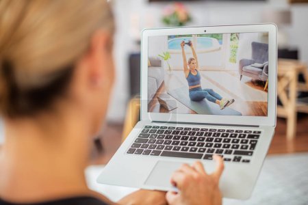Téléchargez les photos : Vue arrière d'une femme pratiquant un entraînement de musculation à la maison avec une leçon vidéo sur ordinateur portable pendant la journée. - en image libre de droit