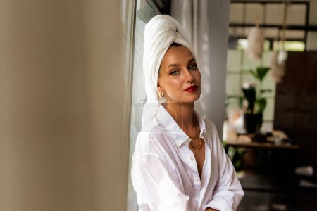 Téléchargez les photos : Portrait de magnifique jeune femme souriante et portant une chemise blanche et une serviette turban sur la tête tout en se relaxant à l'intérieur. - en image libre de droit