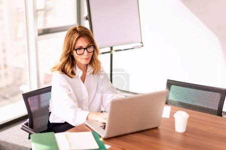 Téléchargez les photos : Femme professionnelle d'âge moyen portant une chemise et utilisant un ordinateur portable tout en travaillant dans un bureau moderne. Femme d'affaires attrayante assise au bureau. - en image libre de droit