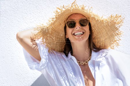 Foto de Primer plano de una atractiva mujer con sombrero de sol y gafas de sol mientras está de pie en la pared blanca. Mujer feliz disfrutando del sol. - Imagen libre de derechos