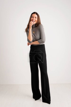 Photo for Full length of an attractive woman toothy smiling and looking at camera. Brunette haired female wearing turtleneck sweater and trousers while standing against isolated white background. Copy space. - Royalty Free Image