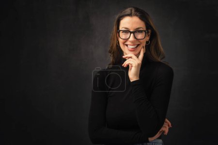Brunette haired woman cheerful smiling and standing at isolated dark bacground. Attractive female wearing turtleneck sweater and eyewear. Hand on chin. Poster 653022960