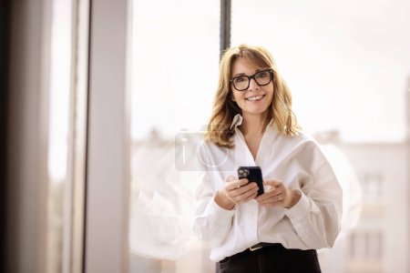 Foto de Retrato de una mujer de negocios de mediana edad de pie en una oficina moderna y mensajes de texto. Mujer de pelo rubio con gafas y camisa blanca. - Imagen libre de derechos