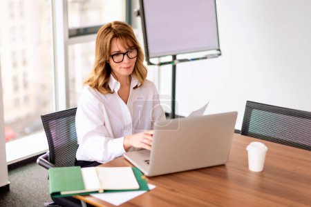Photo for Mid aged professional woman wearing shirt and using laptop while working in a modern office. Blond haired businesswoman wearing eyewear. - Royalty Free Image