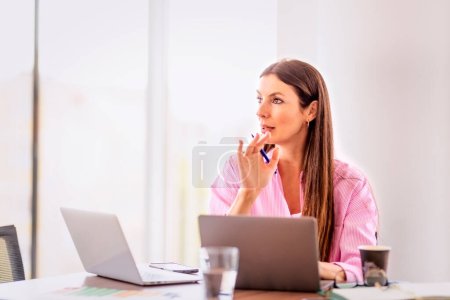 Foto de Pensando en la mujer de negocios usando computadoras portátiles mientras está sentado en el escritorio y trabajando en una oficina moderna. - Imagen libre de derechos