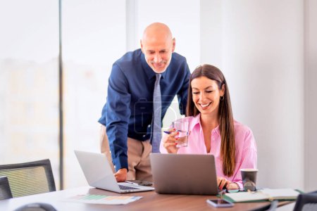 Photo for Group of business people having a business meeting. Attractive female sitting at the table in conference room and professional man standing next to her and working on laptops together. Teamwork. - Royalty Free Image